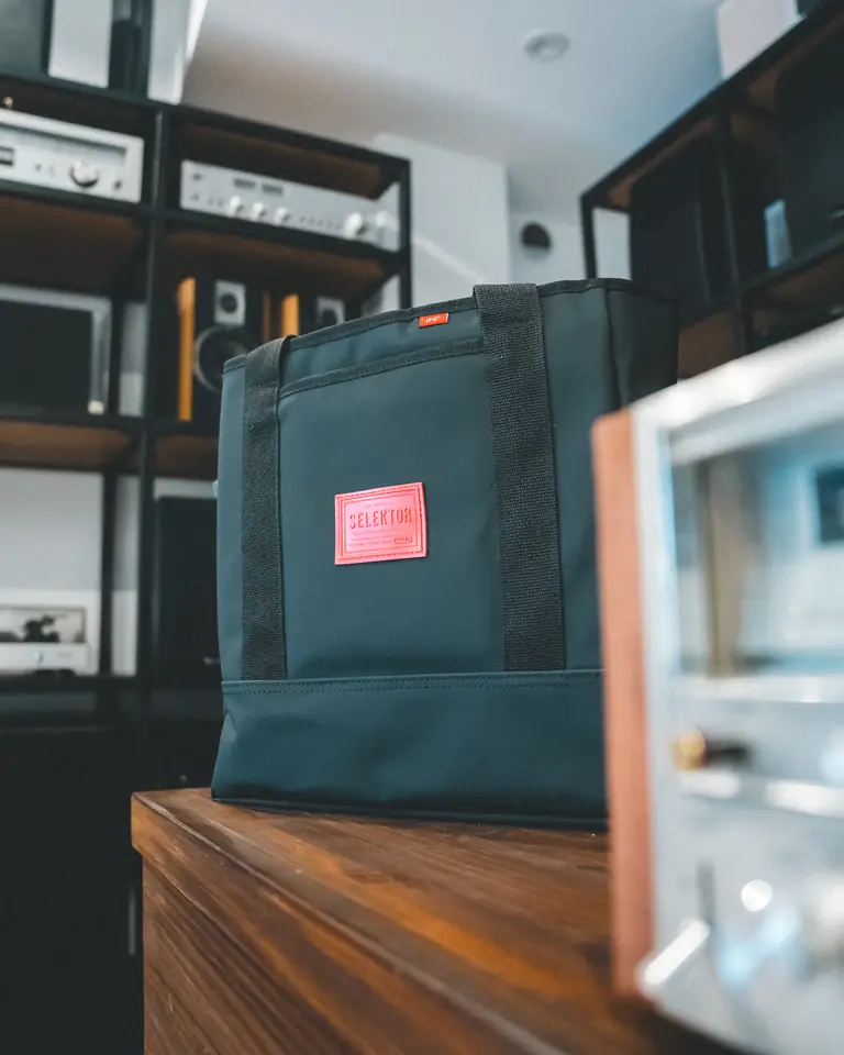 Bolso Selektor verde petróleo con parche rojo, apoyado sobre un mueble de madera junto a un equipo de audio vintage